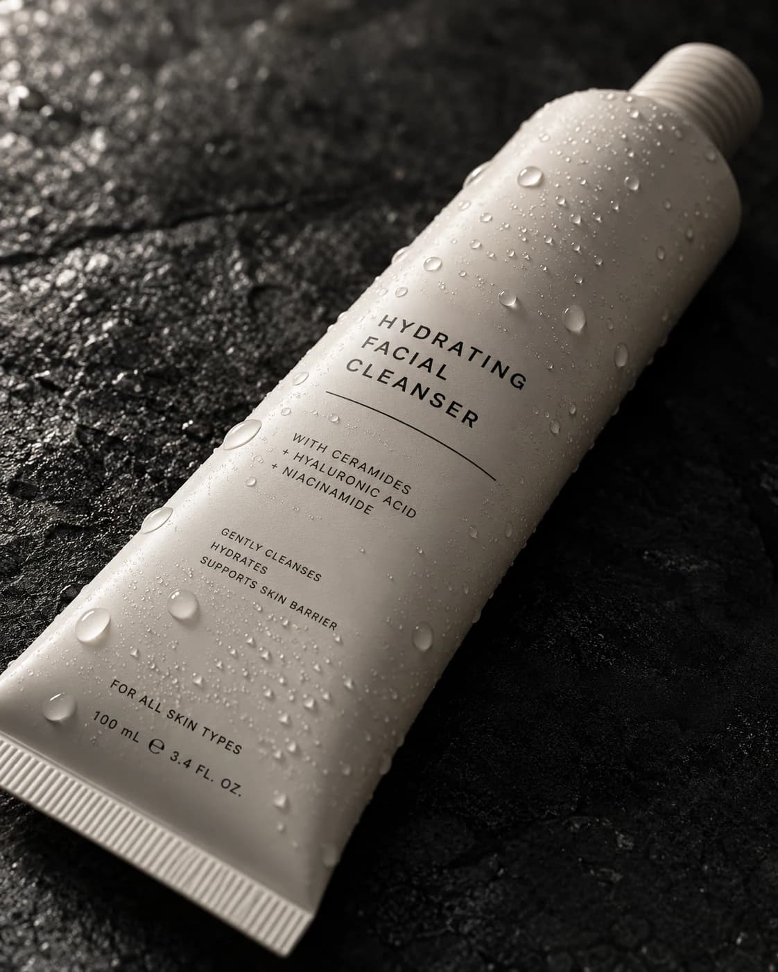 Macro product render of a matte off-white cosmetic tube on dark stone, with water droplets across the packaging.